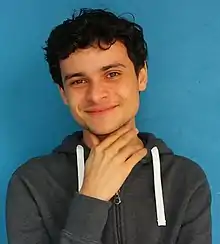 Brunnet man with a smile with his hand close to his neck wearing a jacket stand over a blue background