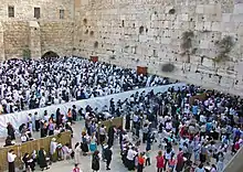 Two large groups of people, seen from slightly above them, separated by a white cloth barrier, standing before a beige stone wall whose top cannot be seen, with another wall in the rear. The group in the foreground is all female, the one in the rear is all male, with many wearing white robes or shrouds