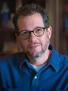 A photo of Michael Giacchiano in 2017.  He's sitting against a blurred background wearing a light blue button-up.