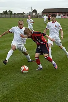 F.C. United players challenge a Mickleover Sports player for possession of the ball while a crowd of supporters watch from the far end of the pitch near the Mickleover goal.