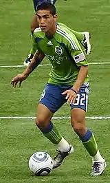 Two men in blue and green soccer uniforms. One has the ball at his feet and is looking upfield.