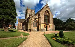 A Hamstone (yellow) building with a central tower. Closest to the camera is the end of the building with a stained glass window. The foreground shows paths and lawns.