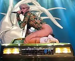 A short-haired blonde woman dressed in a marijuana-themed leotard is kneeling on a green sport utility vehicle with a microphone held to her mouth.