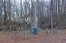 A group of graves inside a small metal railing low to the ground. In the middle is a large gray obelisk. Behind are bare trees, through which a nearby yellow house is visible.