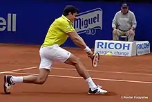 Raonic sliding forward on clay, leading with his right leg. He is about to hit a two-handed backhand shot. In the background, a line judge sits.