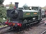 A pannier tank locomotive is passing a station platform, running round its train which includes rolling stock visible in the background. The pannier tanker is green, apart from the black chimney, brass safety valve cover,  red buffer beam, and grey coupling rods. Great Western is written on the side of the tank, and 7752 on a number plate on the cab