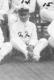 A man sitting cross-legged on a wooden deck, wearing a plain white suit and a white peaked cap with black visor. The number "22" is written over his image, and around him are other seated men with numbers similarly written over them.