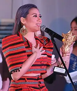 Morissette, in a vibrant red and black striped outfit, delivering a speech into a microphone while holding a trophy