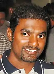 A smiling, dark-skinned man with short black hair and trimmed beard. Several people are in the background.