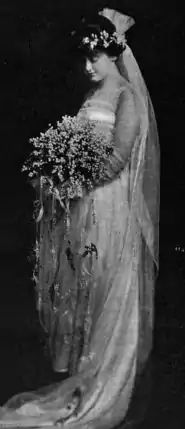 A young white woman standing in a wedding gown, holding a large bouquet.