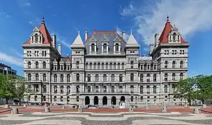 An ornate building, several stories high, of light colored stone. Many arches are visible on its front. On its sides are two large towers with pyramidal red roofs, echoed by similar smaller towers closer to the center with stone tops. In front of the camera, at bottom, is a plaza with a wavy-line pattern