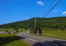 A two-lane paved road winding through countryside from just right of the camera, down the center of the frame, towards a hill covered with green trees under a blue sky with some small clouds in it. On the far side of the road there is a sign with the number 22 on it; below it is a white on blue sign with "Be Prepared to Stop" on it in capital letters. Telephone wires enter the image from top left, connecting to a wooden pole at the center