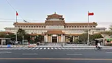 An ornate building front stretching the width of the image, slightly tilted, against a uniformly blue sky. Its front has a projecting pavilion in the Chinese style, echoed by a similar pagoda-style top on the roof above it.