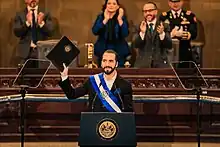 A man (Nayib Bukele) standing at a podium and raising a legislative proposal in his right arm.