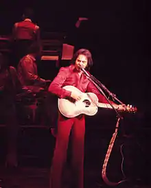 A long-haired man wearing a purple shirt, playing a guitar and singing into a microphone in front of other musicians