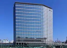 A modern multistorey building in blue and grey colour, with Japan Airlines' "JAL" logo on the top right, blue sky on the background, and a highway bridge in the foreground