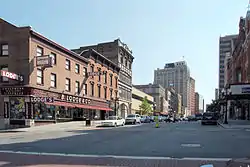 A view down an urban street in a well-developed area. There are lower buildings in the foreground, including one with "B. Lodge & Co." prominently displayed on it at the left, across the intersection from the camera. In the rear are taller ones.