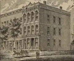 A sepia-tone illustration of the Northampton Bank in 1876. It is a three-story rectangular building, visible here on two sides. Windows are on both sides of the building on all three floors. The phrases "Savings Bank" and "Northampton Bank" are on both sides. It is adjacent to another building on its left, and a dirt path goes past its right. In front, two people are walking, and one person is on a horse-drawn carriage.