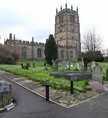 A church seen from the northeast, the tower to the right; both the tower and the body of the church have crenellations and pinnacles. In front of the church is the churchyard with a variety of gravestones