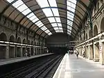 A railway station with side platforms either side of two tracks that disappear into darkness under a painted steel bridge like structure topped with a brick wall, covered by a partially glazed barrel roof.