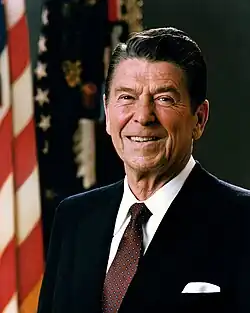 A man in his 70s, with black gelled hair parted on the right, wearing a dark suit and spotted tie, smiling; flags, including the Stars and Stripes, in the background