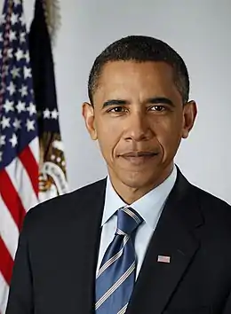 A man wearing a black suit and blue-striped tie, with the American flag in the background