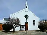 Straight-sided gable end with small belfry at the apex, a rosette window and arched windows and door. Wall is moulded along upper edge and has heavy moulded corner quoins. Arched 3 × 3 panel double door. 2 high, arched, small-pane windows (18 + 4 panes Type of site: Church Current use: Theatre.