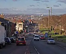 Ina suburban area, a busy main road with vehicles travelling in both directions recedes into the distance. On the left is a row of red-bricked two-storey houses. On the right, an open parkland fringed with mature trees is visible. In the distance is a sunlit landscape of urban and industrial buildings leading to distant, low hills.