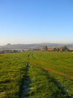 A lightly rutted field in winter, with several buildings in the middle distance and hills behind. The buildings are partly obscured by fences, and consist of farm outbuildings, a large farmhouse with a red-tiled roof and chimneys, a smaller adjacent building in a similar style, and a two-storey house.