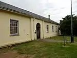 Perimeter wall around three sides. Front range with pediment missing. Slate roof to front. Corrugated elsewhere. Brickword plastered. Mainly single storeyed. Centre block double storey. The construction of this gaol commenced on 16 April 1813. In April 1822 the Magistrate of Albany reported that the gaol was too small, and was offensive in that a residential area adjoined it. It was [closed?] in 1824 whereafter it became a school and later a library. Type of site: Gaol Previous use: Prison. Current use: Offices.