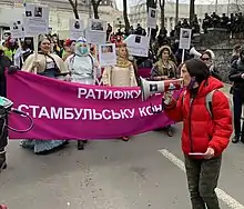 Photograph of a woman in a red coat using a bullhorn to urge on women marchers who are carrying a banner.