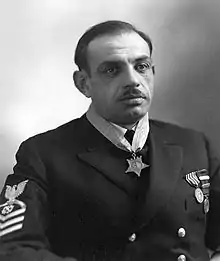 Head and torso of a man with a mustache and dark, thinning hair wearing a dark jacket with stripes on the upper sleeve, two round medals on the left breast, and a star-shaped medal hanging from a ribbon around his neck.