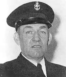 Head of a man wearing a dark jacket and tie and a peaked cap with an anchor and "USN" emblem on the front.