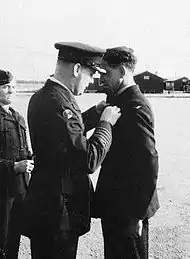 Three men in dark military uniforms; one pins a badge on another's chest