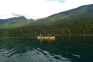 Two paddlers sit in a canoe, gently paddling across the calm water. Behind them, large green mountains flank the water.