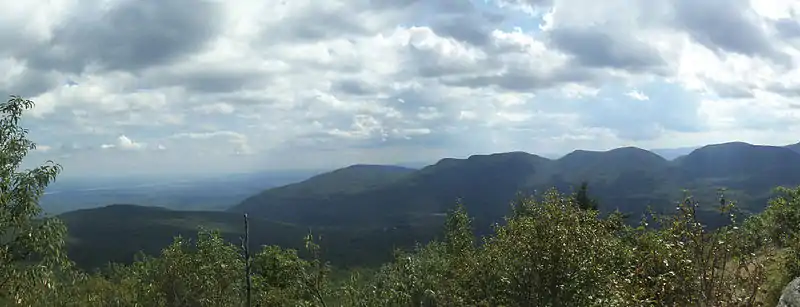 A view over short trees to distant mountains, dappled by intermittent sun and shadow. To the left a river flows through a much lower landscape