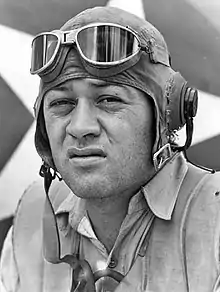 Head of a squinting man wearing a shirt unbuttoned at the collar and a cloth aviator's cap with headphones built into the ear flaps, an unbuckled chin strap, and goggles pushed up onto his forehead.