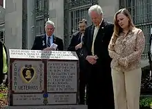 A color picture of Daniel and Maureen Murphy standing next to a monument in front of the Lieutenant Michael P. Murphy Post Office in Patchogue, New York. The monument has a purple heart and some wording inscribed on it and there are two men in the background.