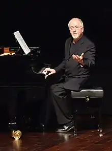 Patrick Doyle sitting at a grand piano in Quitar in 2016. He is sitting with one hand on the keys, and the other hand gesturing while facing the audience.