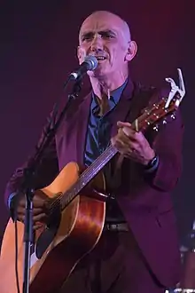 Head and shoulders of a sixty-year-old male standing at a microphone. He is strumming a guitar, singing and staring into the distance. He wears a purple suit, tie, blue shirt and vest.