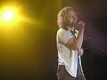 A male singer, Eddie Vedder, onstage and singing into a vocal microphone. He has an emotional look on his face as he sings.