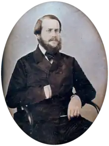 Photographic half-length portrait of a seated bearded man dressed in a dark, double-breasted coat with his right hand tucked inside the front