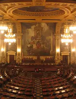 A view of a large, ornately-decorated room with several rows of curved desks arranged in a semicircle. A large mural is visible on the wall at the far end of the room.