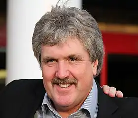 A headshot of a grey haired Caucasian man with a moustache wearing a suit.