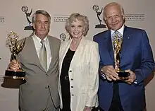 Three people standing in front of a banner are smiling at the camera. Two men are holding Emmy statuette with a woman in the middle