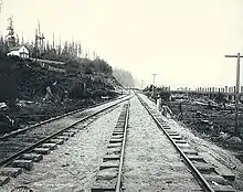 A black-and-white photograph of two railroad tracks against an undeveloped hillside with trees and a small house.