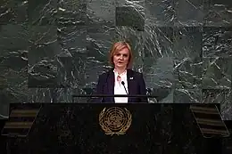 Truss, wearing a white shirt and purple coat, speaks at a podium in front of a greyish-green marble wall.