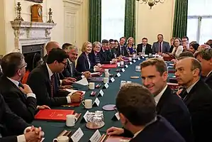 A room full of people sitting in a white room at a dark green table. Truss is roughly in the centre-left of the image, smiling.