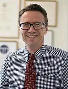 middle-aged white male with short brown hair, wearing glasses, a dark red patterned tie, and a blue-and-white checkered shirt, grinning at camera