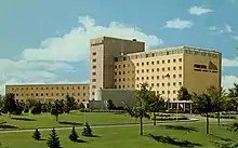 A photo of a beige building in a 1960s modern architecture style stands before a manicured green lawn upon which stand several low trees and shrubs. The building has two wings, the left of which is three stories tall and the right of which is seven or eight stories high, with nine-story tower connecting them. The sky is blue with some clouds.